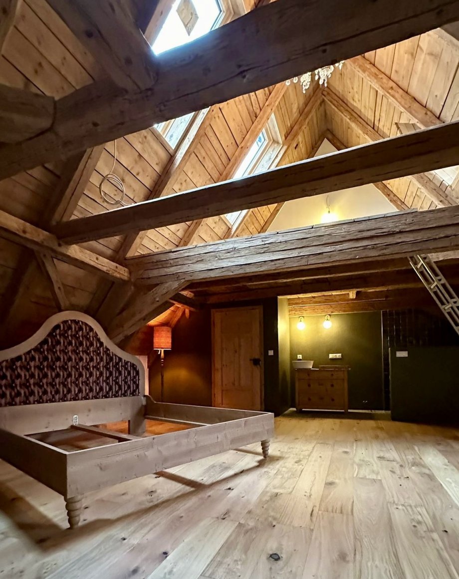 Op maat gemaakt bed met gebogen hoofdbord onder een monumentale houten balkenkap, met dakramen en groene wand op de achtergrond, door cestsjoukje