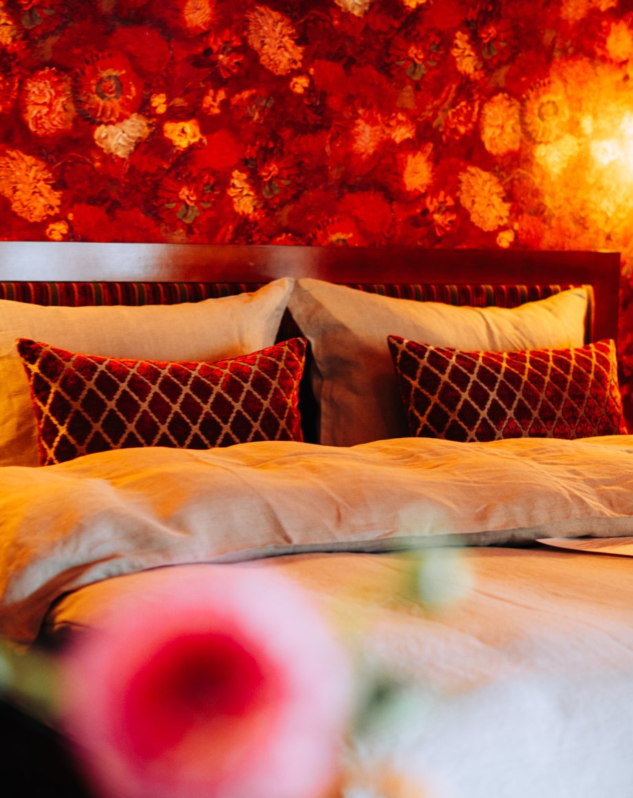 Hotelkamer met warm rood bloemenbehang, fluwelen ruitkussens en zandkleurig beddengoed, door cestsjoukje