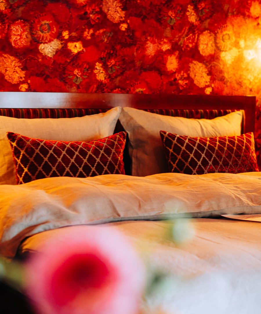 Hotelkamer met warm rood bloemenbehang, fluwelen ruitkussens en zandkleurig beddengoed, door cestsjoukje