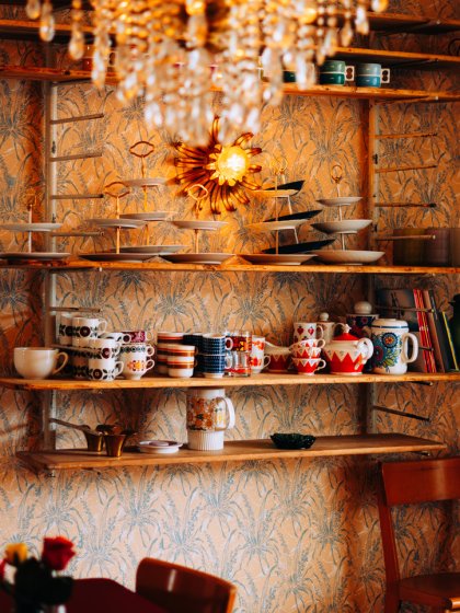 Cafe-interieur met open wandrek vol bont retroservies, etagères en kristallen kroonluchter tegen botanisch behang, door cestsjoukje